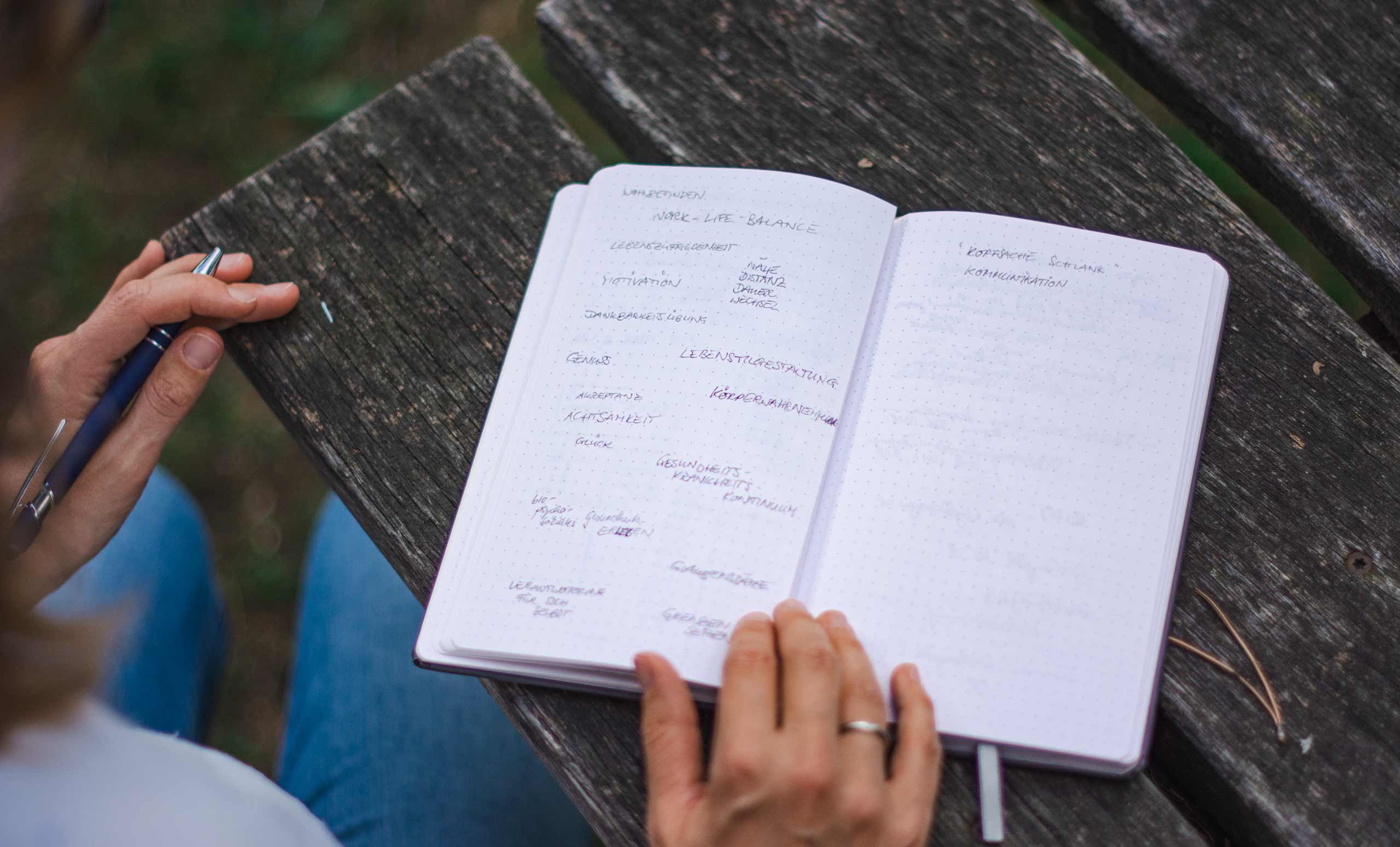 Hand hält Stift, im Buch daneben einige Notizen mit Schlagwortern zum Thema Gesundheitspsychologie